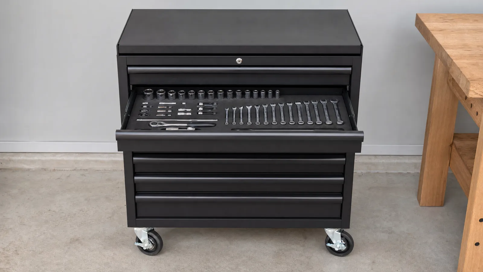 A black rolling tool chest on a concrete garage floor with one drawer open, showing sockets and combination spanners laid out in black foam