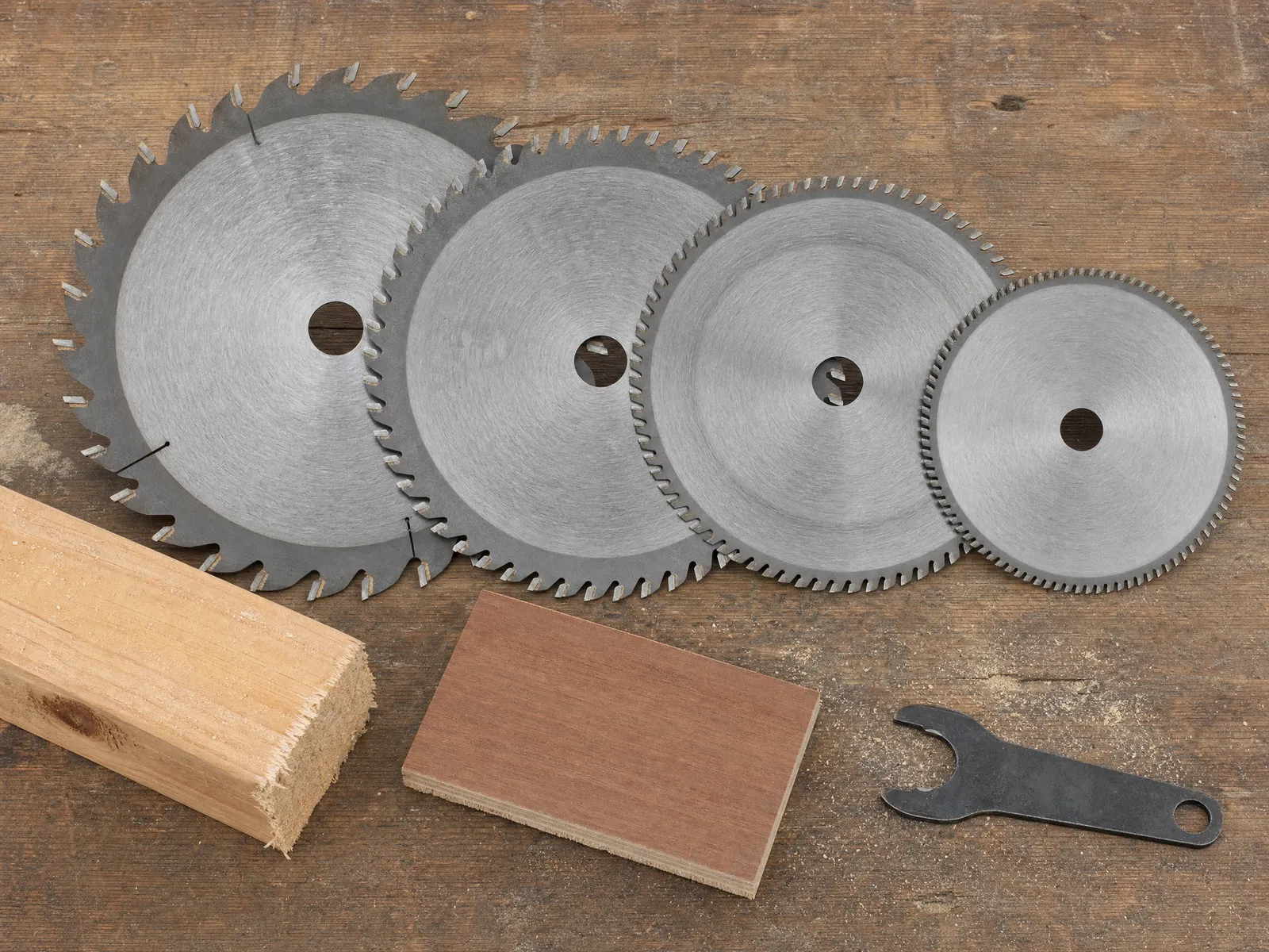 Four circular saw blades laid out from coarse to fine tooth count on a workbench beside a rough-sawn framing offcut and a clean-cut plywood offcut